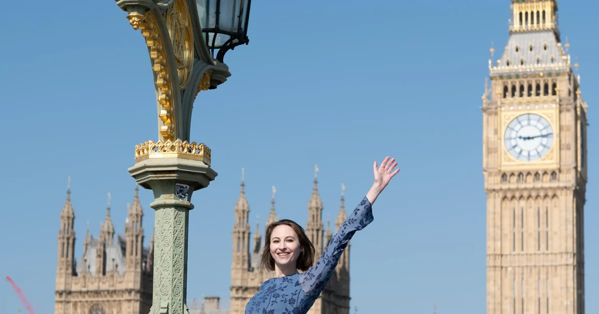 A woman in a blue dress dancing by a lamp post on Westminster Bridge with Big Ben behind her