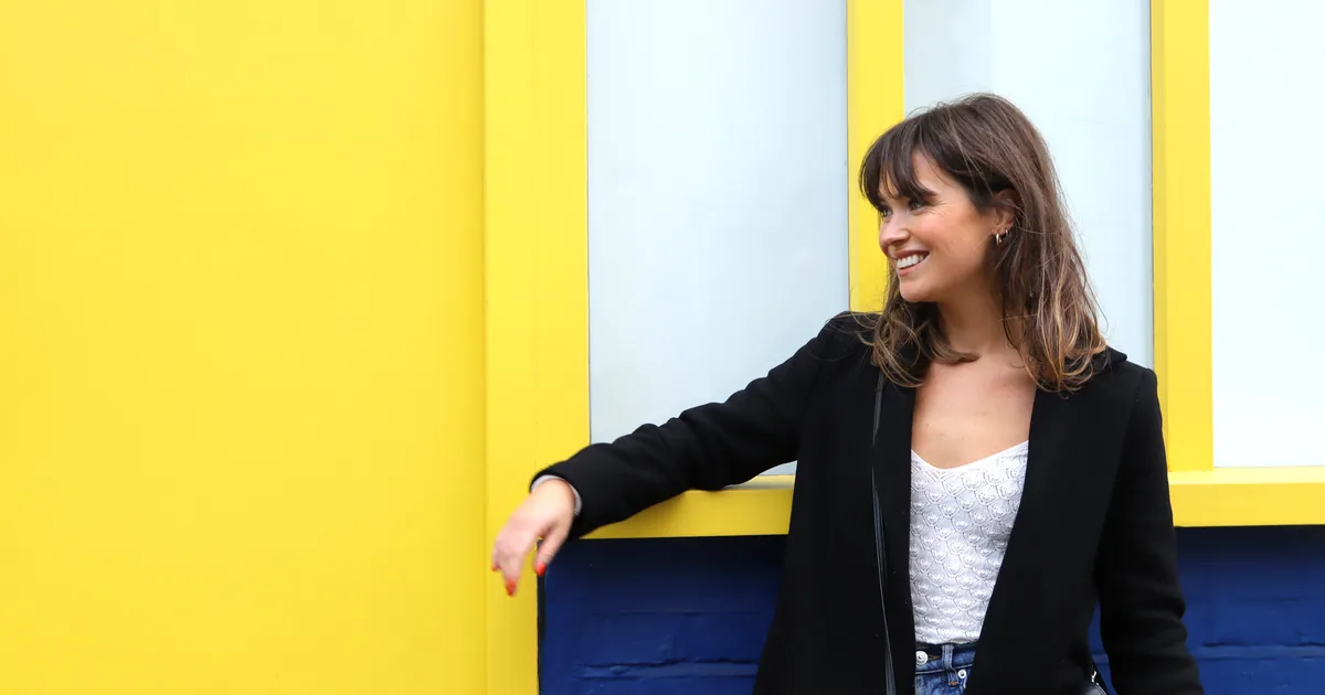 A woman in a black jacket and white top leaning against a bright yellow wall in Notting Hill